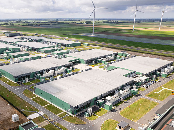 Aerial view of a large area of land with multiple warehouse type buildings, a water tank, and roads with wind turbines and farm land in the background