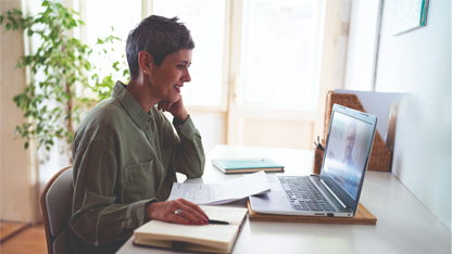 A woman in a green top sits at a laptop in a bright room. 