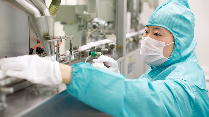 A pharmaceutical factory worker wearing a mask and protective attire is engaged in machine operation, highlighting workplace safety measures.