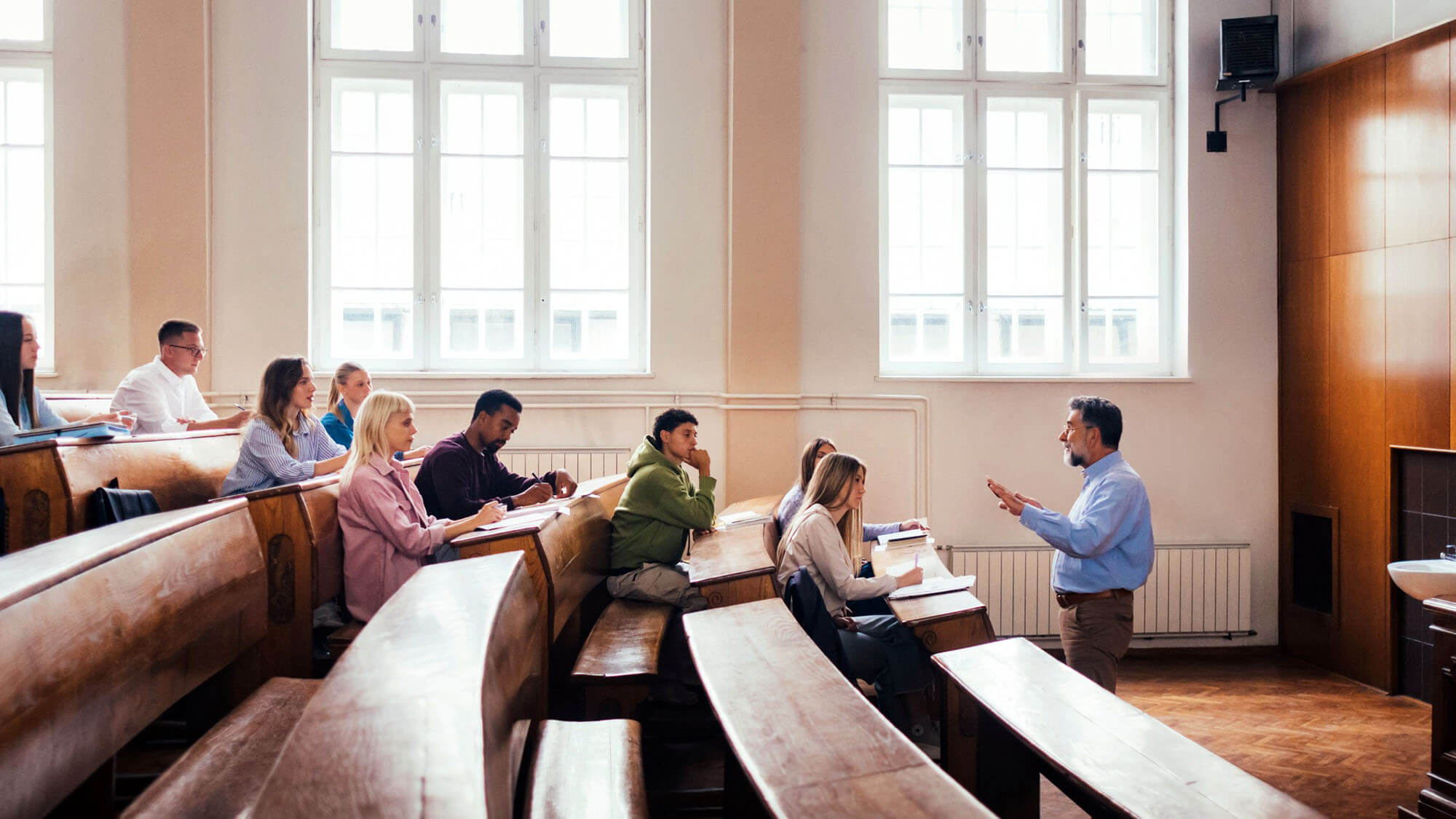 A professor teaches a group of attentive students in a university lecture hall. The classroom setting fosters an engaging educational environment with diverse individuals and learning activities.