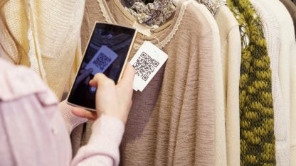 A person uses a smartphone to scan a QR code on a clothing tag while browsing sweaters on a store rack.